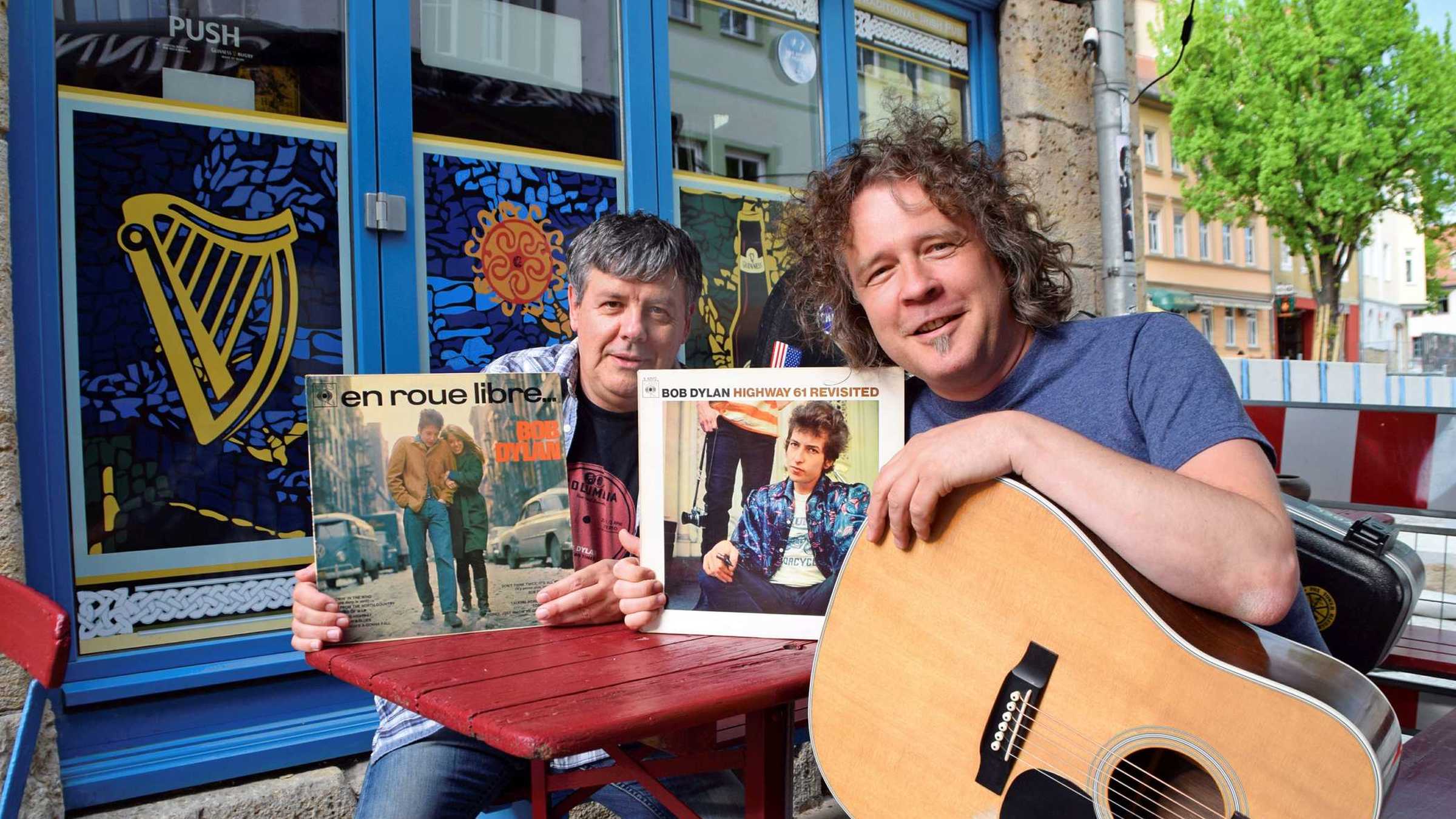 Die zwei Mitglieder der Band halten Bob Dylan Platten in die Kamera. Einer hat eine Gitarre auf dem Schoß