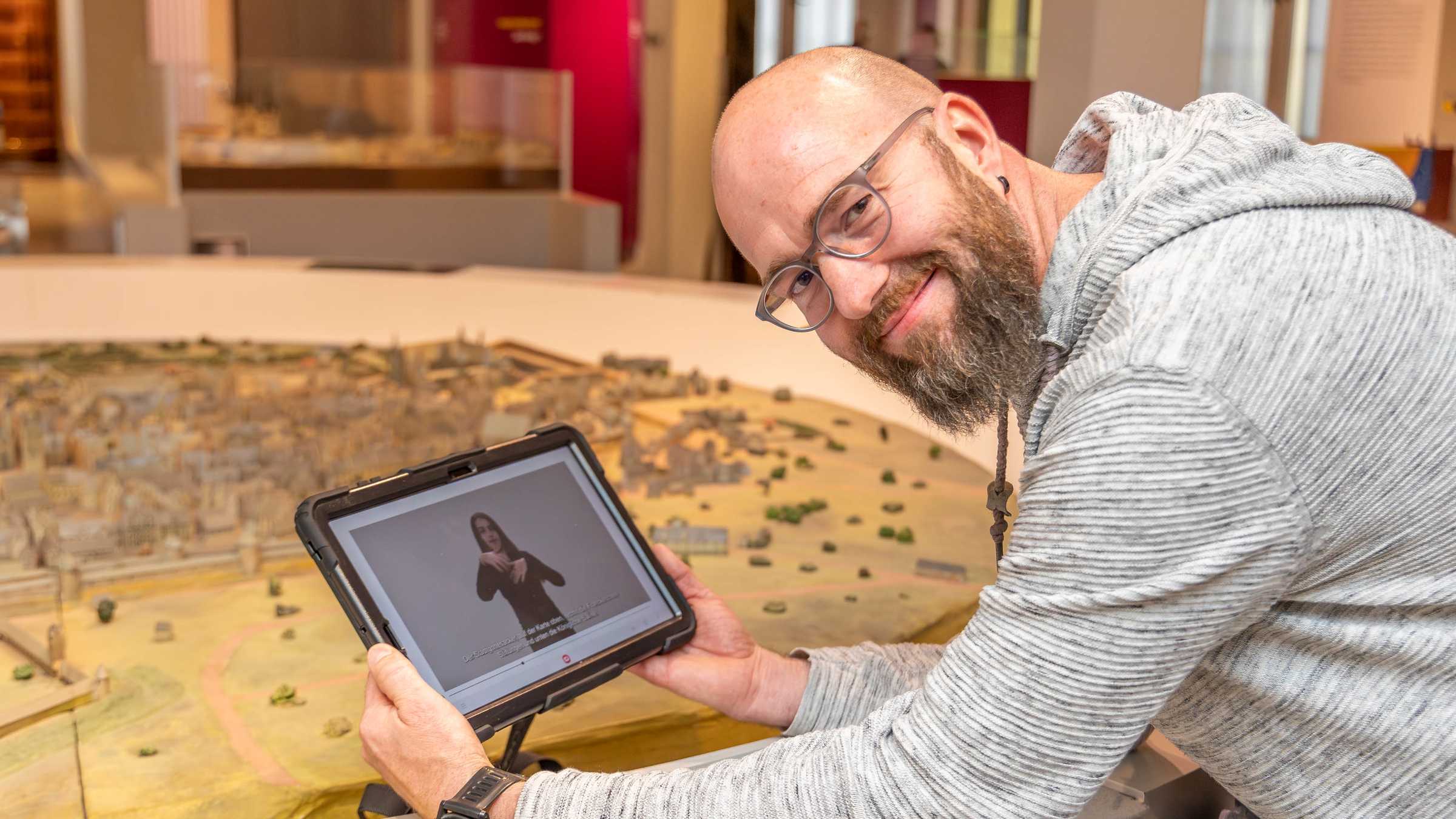 Henry Niekrawietz mit App Henry Niekrawietz mit einem Tablet, auf dem eine DGS-Führung gezeigt wird, im Stadtmuseum vor einem Stadtmodell