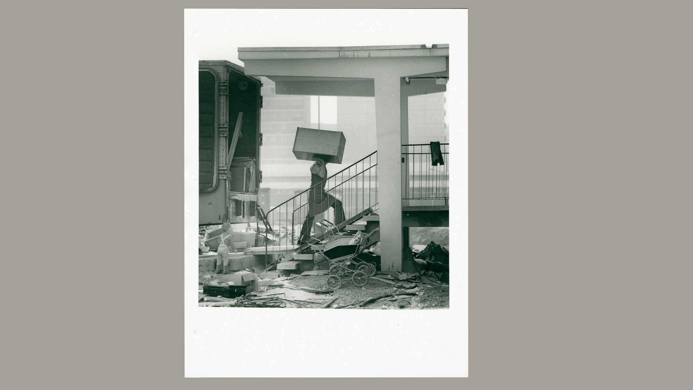 Einzug 033 Halle-Neustadt, Foto: Gerald Große Foto auf dem ein Mann einen Schrank auf der Schulter eine Eingangstreppe beherzt hochträgt. Neben der Treppe parken im noch umherliegenden Baustellenunrat 2 Kinderwagen