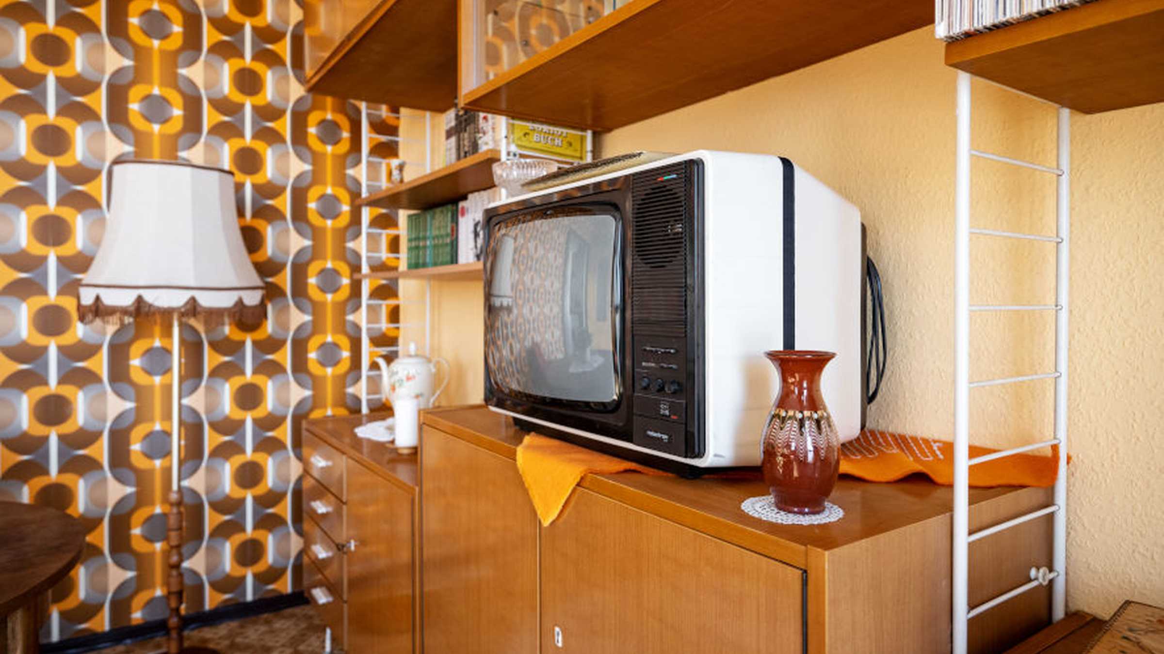 DDR-Wohnzimmer Blick in eine Wohnzimmerecke mit Schrankwand, weißem Kunsstofffernseher, einer braunen Vase und einer groß gemusterten Tapete in Braun-Orangetönen