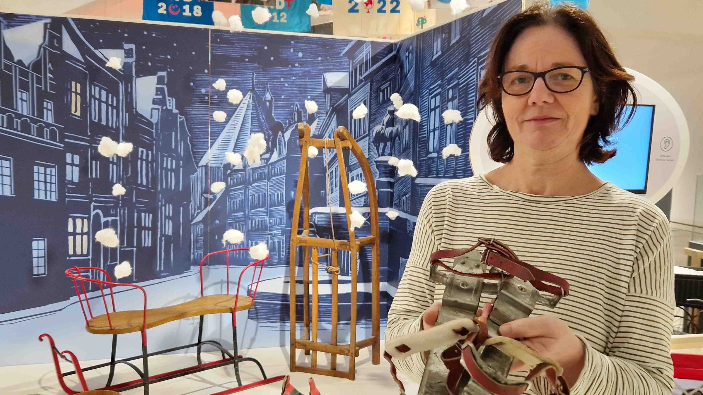 Spiel mit! - Führung mit Ute Fahrig Ute Fahrig hält Gleitschuhe in ihren Händen, dahinter ist eine nachgestellte Winterlandschaft mit Schlitten im Ausstellungsraum zu sehen