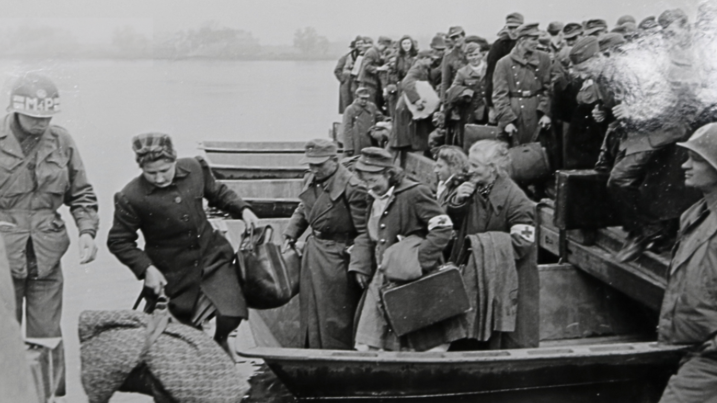 Das Schwarz-Weiß-Bild zeigt eine Gruppe von Flüchtlingen, die aus einem Boot aussteigen. Sie tragen schwere Taschen und Koffer, die sie mit sich führen.