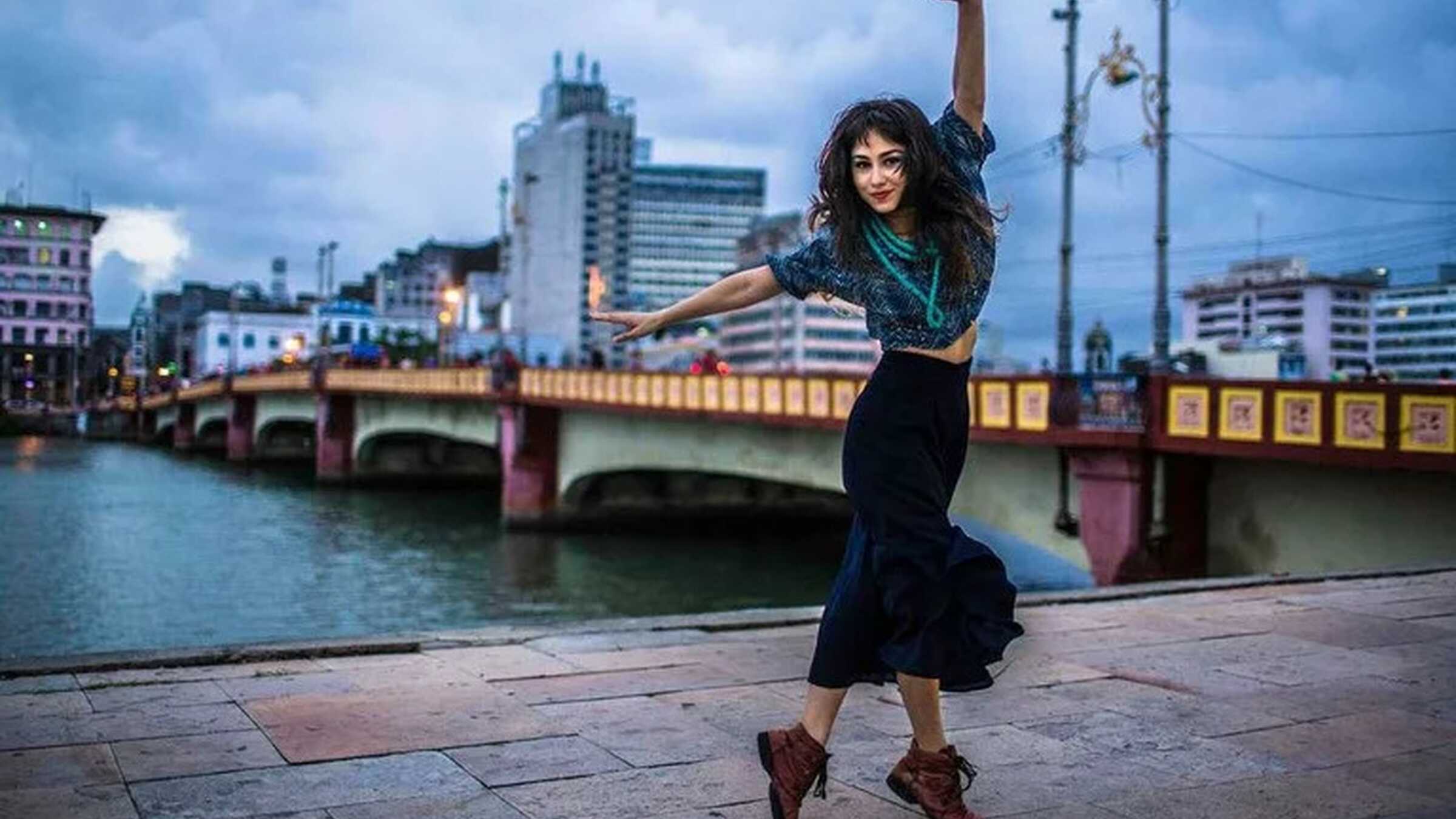 Flaira Ferro in tänzerischer Pose vor einer Brücke
