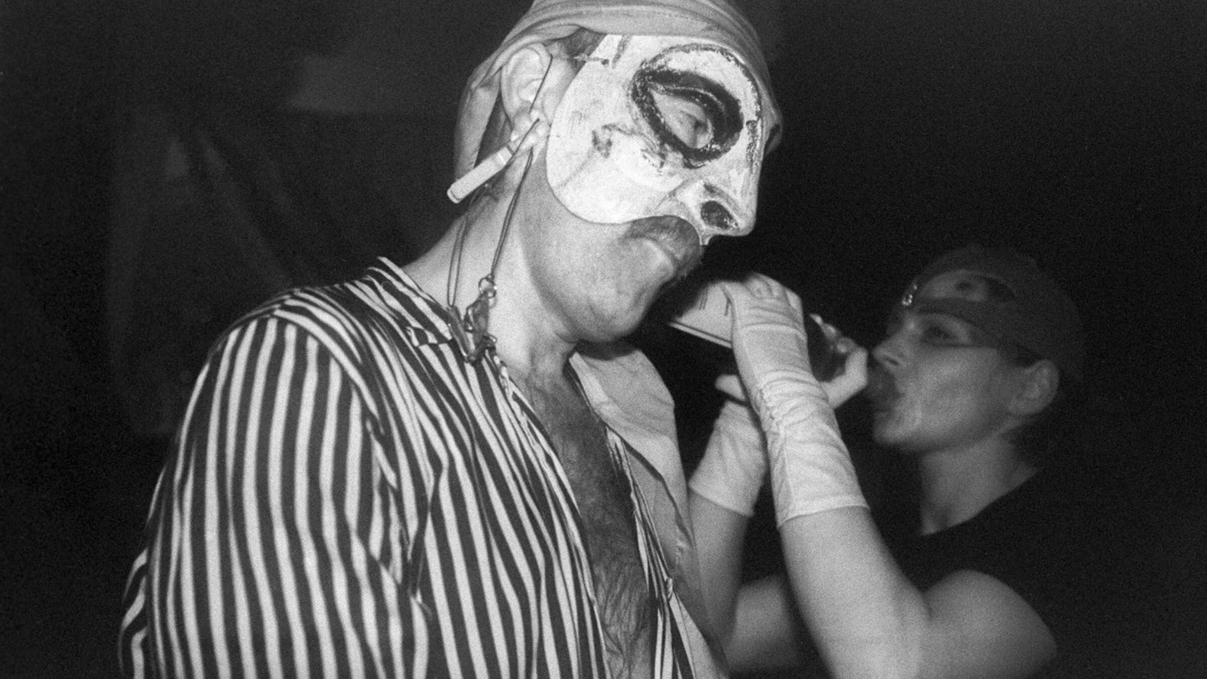 Schwarz-weiß-Foto eines Mannes in gestreiftem Hemd mit Halbmaske, dahinter eine Persin, die eine Flasche mit beiden Händen an den Mund hält