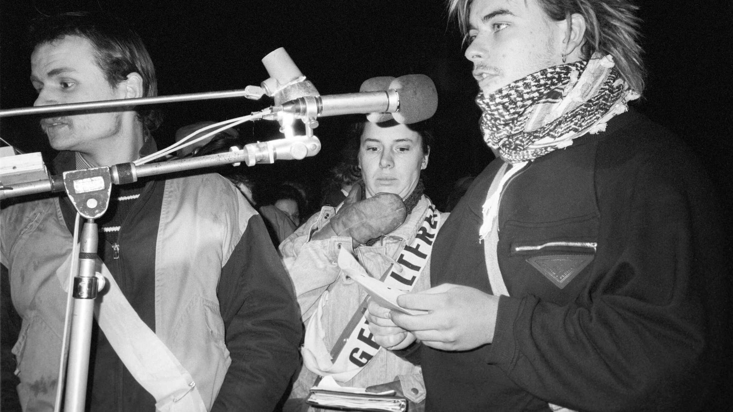 ein junger Mann spricht in ein Mikrofon, dahinter in der Bildmitte steht Thea Ilse mit einer Schärpe, auf der "gewaltfrei" steht