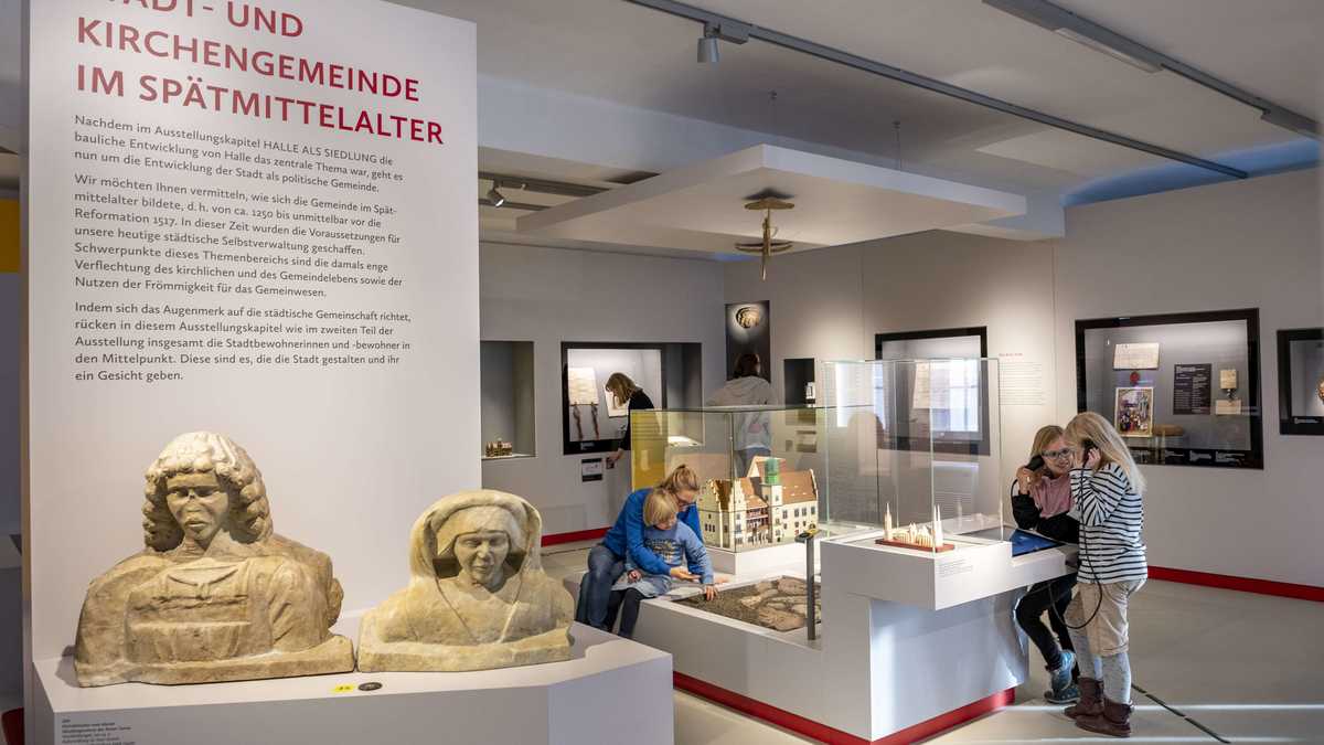 ein großes Ausstellungsschild mit dem Text "Stadt- und Kirchengemeinde". Davor stehen zwei Büsten aus Stein. Im Hintergrund sehen sich Kinder die Ausstellung an