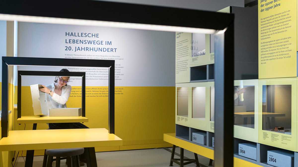 Ausstellung "Hallesche Lebenswege" mit Exponaten auf der rechten Seite und Tischen mit Stühlen auf der linken Seite