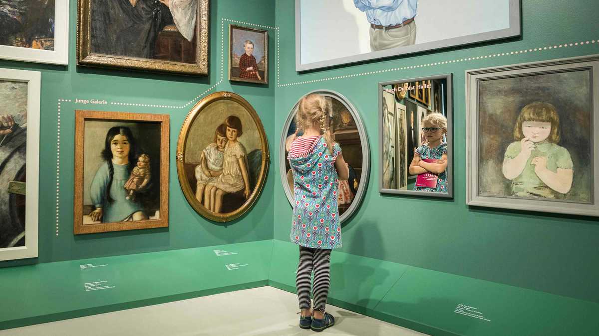 Ein kleines Mädchen in einem Ausstellungsraum "Halle im Portrait". Es schaut sich in einem kleinen Spiegel an