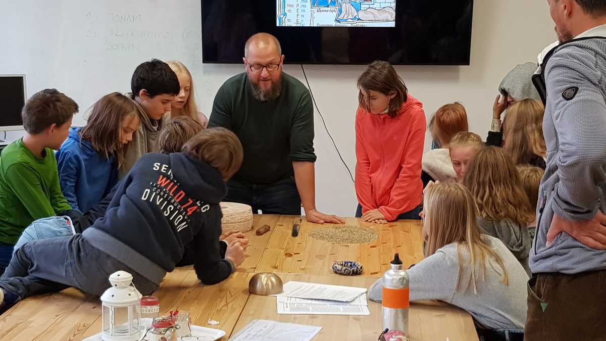 Projektleiter Markus Königsdörfer erklärt den Kindern die mittelalterliche Herstellung von Brot Die Kinder sitzen zusammen mit Projektleiter Markus Königsdörfer an einem Tisch. Im Hintergrund ist auf einem Fernseher eine mittelalterliche Zeichnung zu sehen, auf der ein Mann einen Laib Brot in den Ofen schiebt.