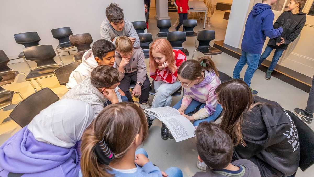 Junge Autor*innen lauschen ihren Geschichten, Foto: Thomas Ziegler Kinder sitzen zusammen und hören einem Mädchen zu, das aus dem Textheft liest