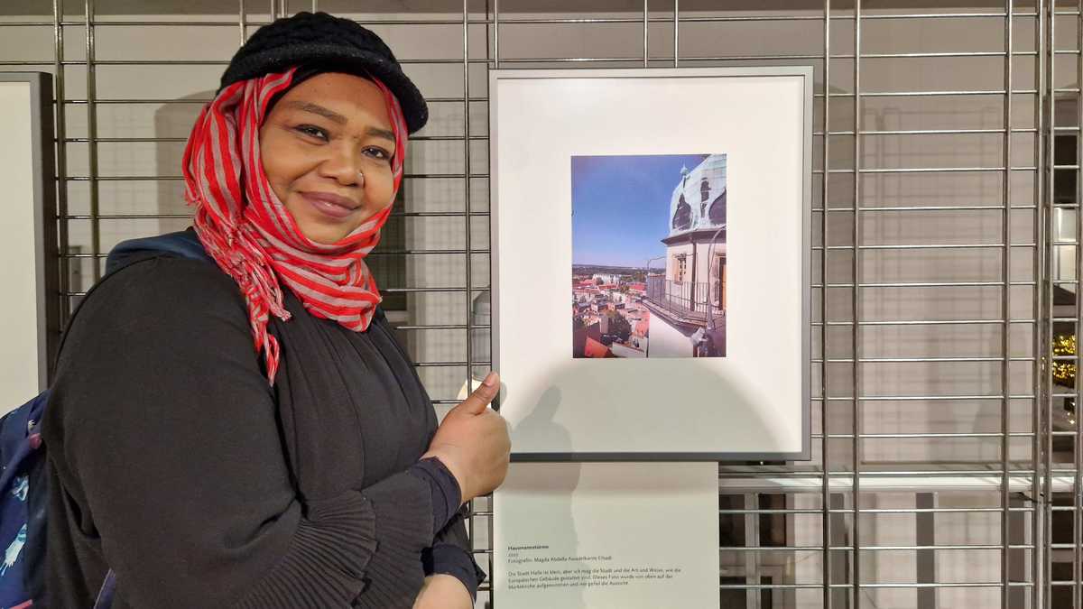 Eine Frau mit dunkler Hautfarbe neben ihrem Foto in der Ausstellung mit Daumen hoch und in die Kamera lächelnd. Das Foto zeigt den Blick von den Hausmannstürmen.