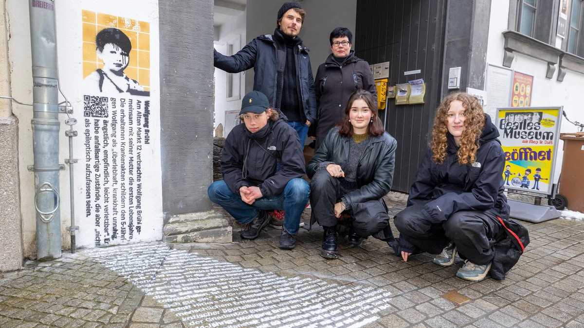 Grafitto am Stolperstein von Wolfgang Brühl 3 Jugendliche aus dem Feiniger-Gymnasium hocken vor dem Grafitto, dahinter Künstler Victor Reichert und Jane Unger aus dem Stadtmuseum