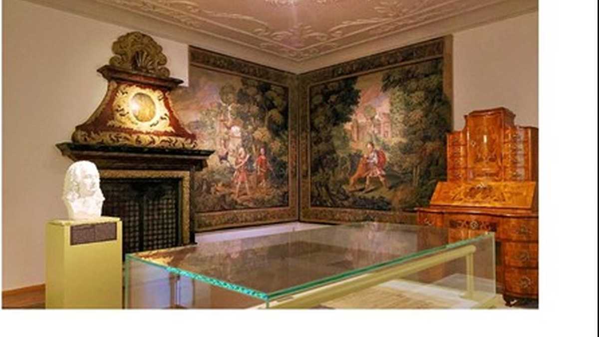 Im hinteren Teil des Raumes sind zwei große Gemälde in der Ecke daneben steht rechts ein Schrank und links ein Kamin im Vordergrund ist eine Glasvitrine und links daneben eine Statue. 