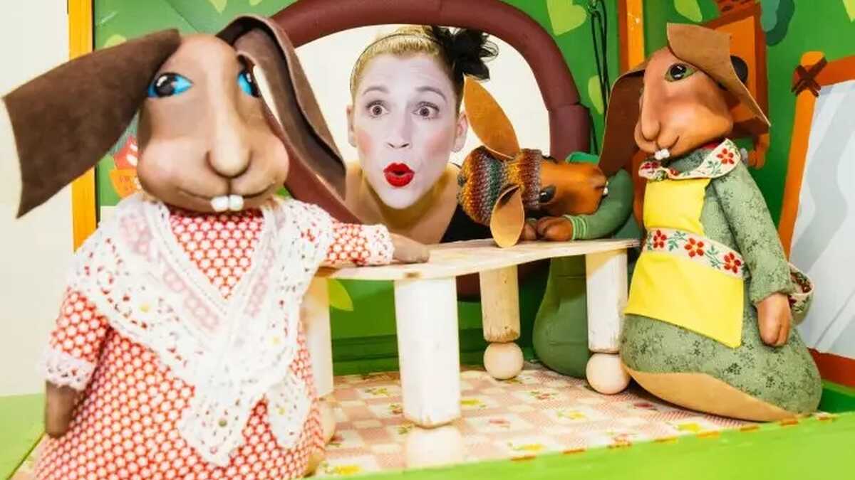 eine Zimmerszene mit Hasenpuppen zu sehen, die Puppenspielerin Julia Raab schaut mit einem überraschten Gesichtsausdruck zu ihnen hinein.