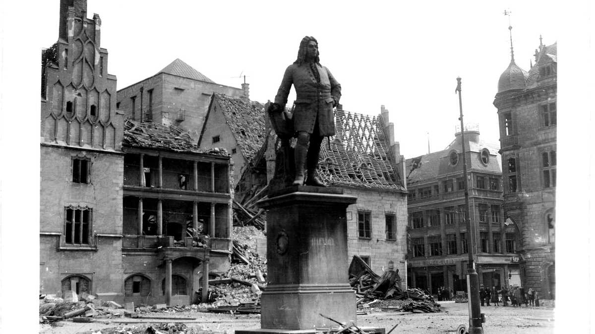 Auf einem städtischen Platz steht eine bronzene Standfigur von Händel auf hohem Sockel, umgeben von Holz- und Bauschutt. Im Hintergrund sind mehrstöckige Gebäude mit zerstörten Dächern und ausgebrannten Fassaden zu sehen; am rechten Bildrand stehen einige Personen.
