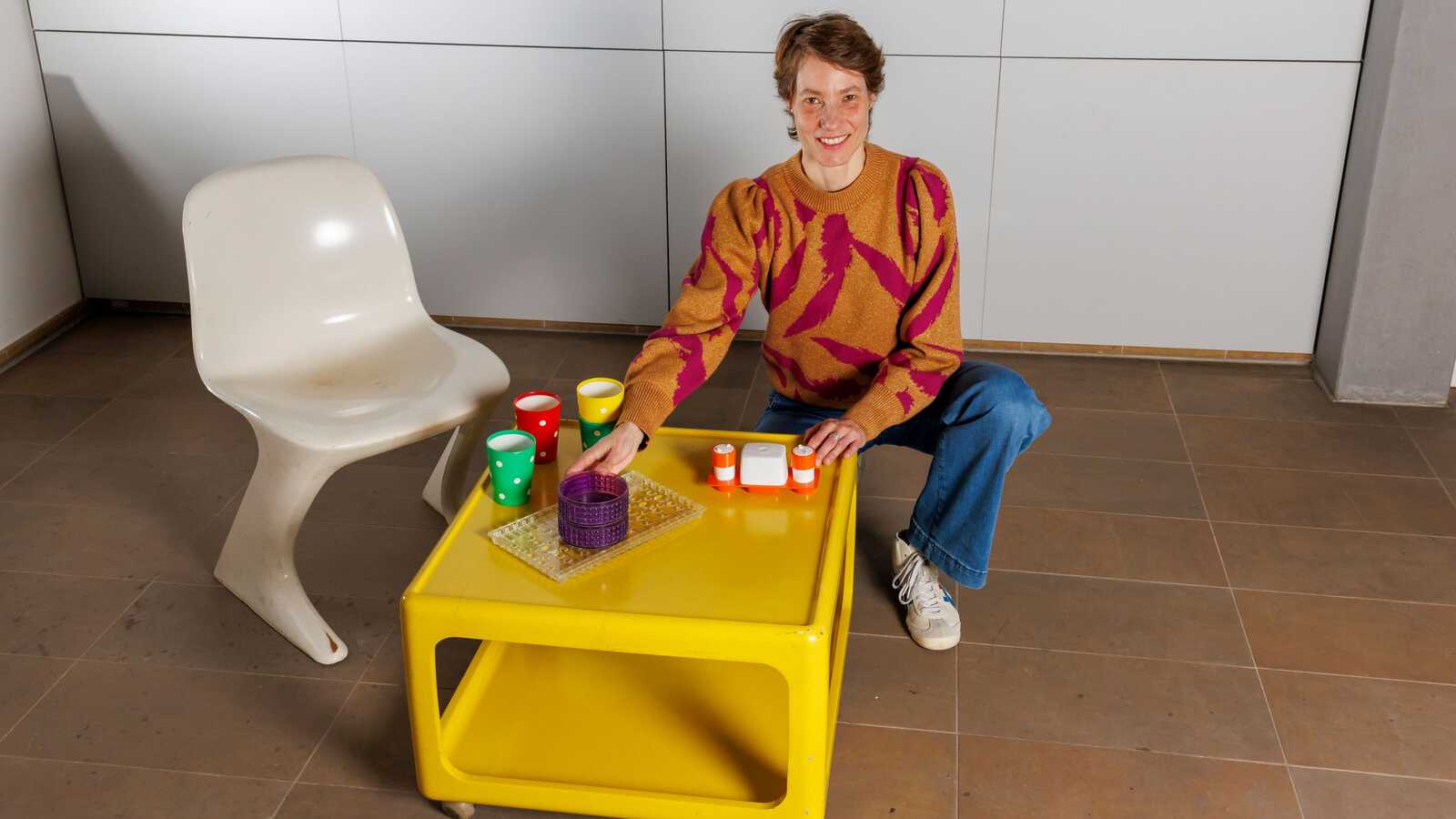 Kuratorin Kristin Land hockend hinter gelbem Couchtisch mit Plastik-Geschirr und weißem Z-Stuhl 