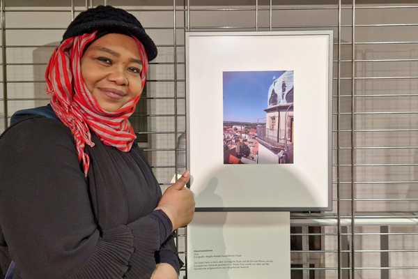 Eine Frau mit dunkler Hautfarbe steht neben ihrem Foto, zeigt den Daumen hoch und lächelt in die Kamera