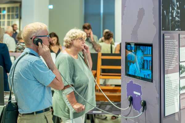 ein älterer Mann und eine ältere Frau mit Kopfhöreren im Ausstellungsraum vor einem Monitor, auf dem ein ein Zeitzeuge erzählt