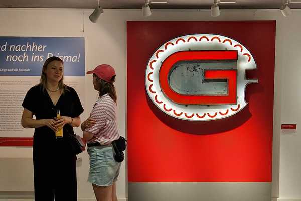 zwei junge Frauen unetrhalten sich in der Ausstellung, im Hintergrund das leuchtende G der Leuchtreklame vom Gastronom