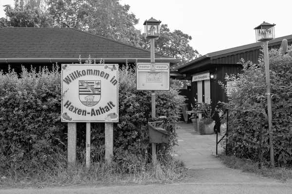 Eine hohe Hecke verdeckt Veraufsbungalows, im Vordergrund ein Schild mit einem Sachsen-Anhalt Wappen mit einem Schwein statt Bär und dem Spruch "Wilkommen in Haxen-Anhalt"