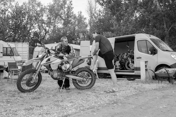2 Junge Männer unterhalten sich an einem Motorrad, im Hintergrund Transporter und Campingstühle