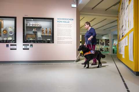 Barrierefreiheit eine Frau begleitet von ihrem schwarzen Blindenhund in der Ausstellung. Diese zeigt die Wahrzeichen von Halle