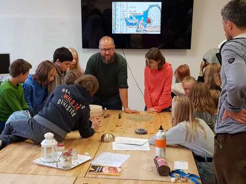 Projektleiter Markus Königsdörfer erklärt den Kindern die mittelalterliche Herstellung von Brot Die Kinder sitzen zusammen mit Projektleiter Markus Königsdörfer an einem Tisch. Im Hintergrund ist auf einem Fernseher eine mittelalterliche Zeichnung zu sehen, auf der ein Mann einen Laib Brot in den Ofen schiebt.