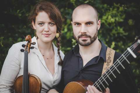 Saela Whim Sarah Graefe mit Geige im Arm und Gianluca Caliva` mit Gitarre stehen vor einem verschwommenen Hintergrund aus grünen Gebüschen.