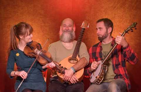 Doc Taylor & The Redhaired Girl Jenny Price, Nico Schneider und Tim "Doc Fritz" sitzen mit ihren Instrumenten vor der Kamera. Hintergrund ist orange-rot.